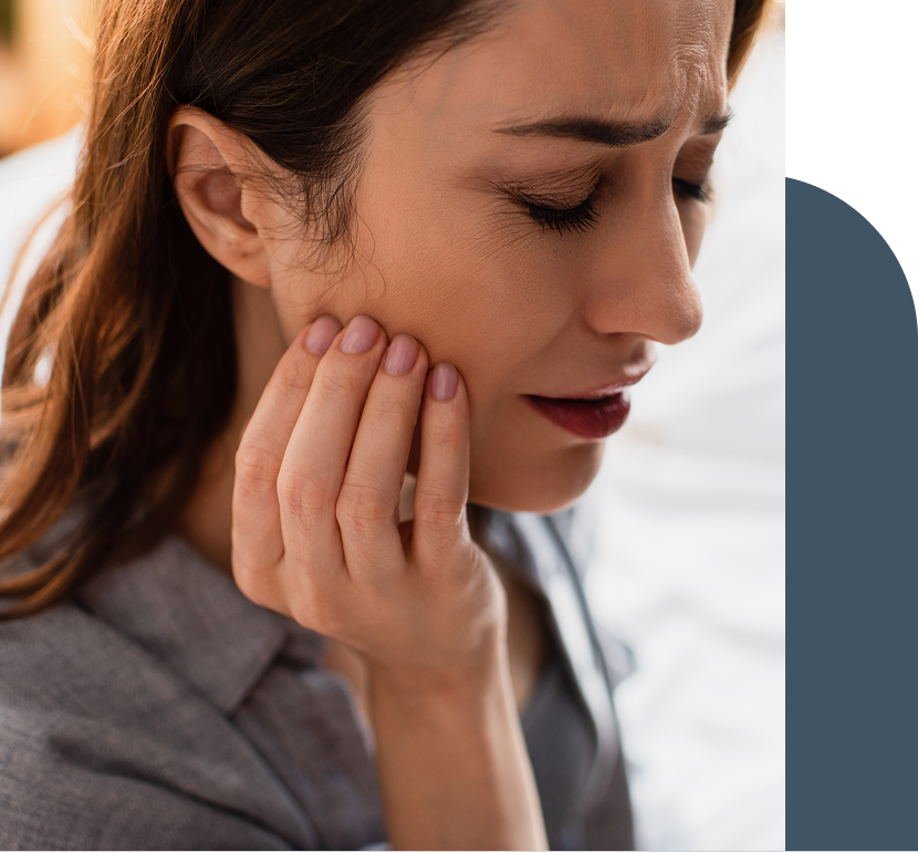 Woman sits with a pained expression, pressing her jaw with her hand, indicating tooth or jaw pain.