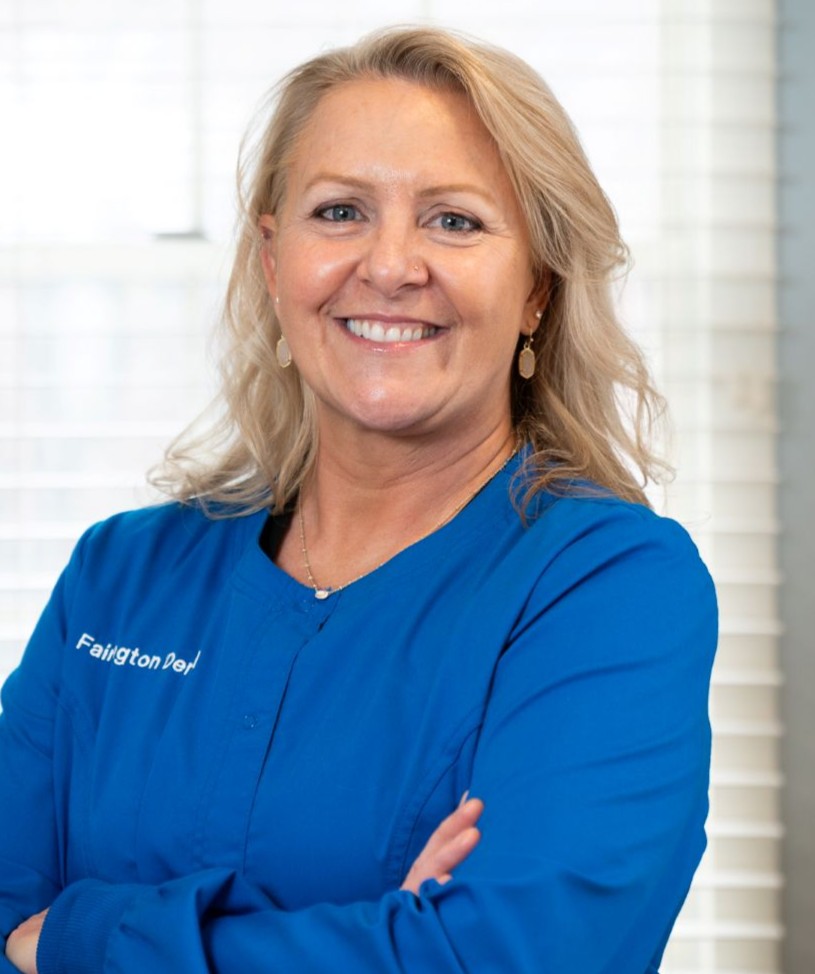 Smiling blonde woman in blue scrubs with arms crossed in a bright clinic.